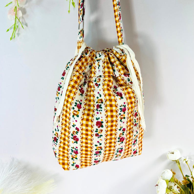 Pode incluir: Uma bolsa com cord&atilde;o com um padr&atilde;o floral e xadrez. A bolsa apresenta uma base xadrez amarela e branca com listras florais verticais em vermelho e verde. A bolsa tem um fecho de cord&atilde;o branco e uma al&ccedil;a a condizer.
