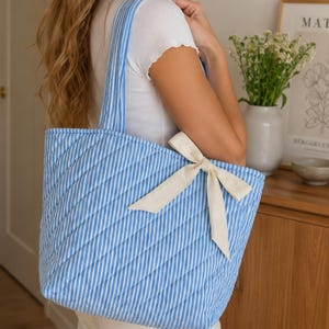 Op de afbeelding: Een gewatteerde tote bag met een verticaal gestreept patroon in lichtblauw en wit. De tas heeft twee bijpassende schouderbanden en is versierd met een crèmekleurig stoffen lint dat tot een strik is geknoopt aan de rechterbovenkant.