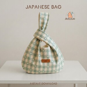 May include: A Japanese bag featuring a light green and cream gingham check pattern with small white hearts throughout. The bag has a knotted handle design and a brown rectangular label on the front with the word HANDMADE printed on it.