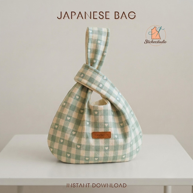 May include: A Japanese bag featuring a light green and cream gingham check pattern with small white hearts throughout. The bag has a knotted handle design and a brown rectangular label on the front with the word HANDMADE printed on it.