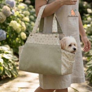 May include: A pet carrier bag featuring a light green and white gingham pattern on the front panel, complemented by a cream-colored fabric with a small floral print on the sides and handles. A small white dog is visible inside the open-top bag.