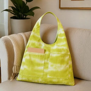 May include: A lime green and white tie-dye hobo bag featuring a single shoulder strap and two large rectangular front pockets. One pocket contains a small, solid tan rectangular notebook. The bag has a triangular shape with a wide base.
