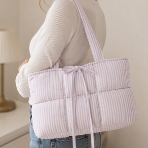 May include: A quilted tote bag featuring a light purple and white vertical striped pattern. The bag has two shoulder straps and a central tie closure made of the same striped fabric. The text "STEP BY STEP GUIDE BEGINNER FRIENDLY" is visible.