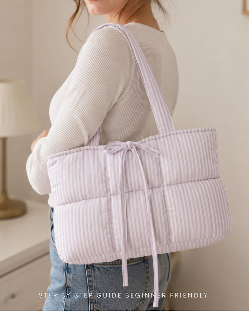 May include: A quilted tote bag featuring a light purple and white vertical striped pattern. The bag has two shoulder straps and a central tie closure made of the same striped fabric. The text "STEP BY STEP GUIDE BEGINNER FRIENDLY" is visible.