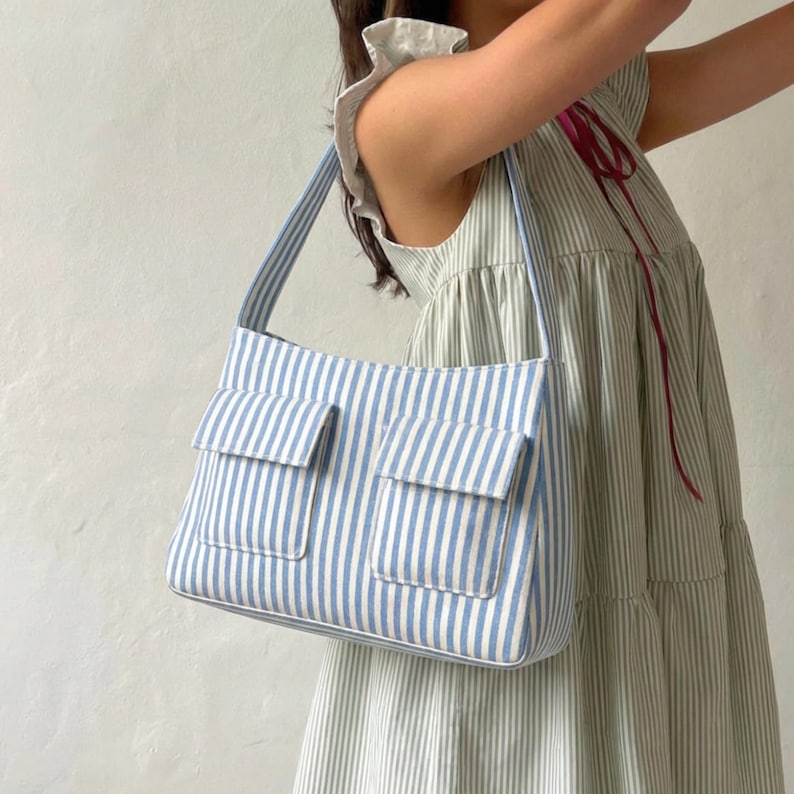 May include: A shoulder bag featuring a light blue and white vertical striped pattern. The bag has two rectangular front pockets with flaps and a single shoulder strap. The fabric is consistent with the light blue and white striped sleeveless dress.