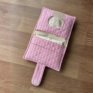 Peut inclure: Une pochette en tissu matelassé à rayures roses et blanches conçue pour contenir des lingettes humides. La pochette présente un rabat rectangulaire avec une ouverture à clapet en plastique, un compartiment central et une longue patte auto-agrippante.