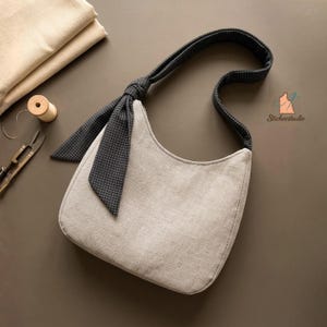 Puede incluir: Un bolso de hombro estilo hobo de color beige hecho de tela similar al lino, con una correa de color gris carbón oscuro con un patrón de cuadrícula blanca fina. La correa está anudada en la parte superior, creando un detalle decorativo.