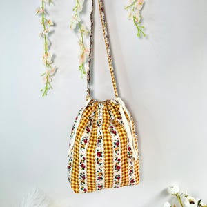 Pode incluir: Uma bolsa de ombro com cord&atilde;o com um padr&atilde;o xadrez amarelo e branco, acentuado com listras florais vermelhas. A bolsa tem um fecho de cord&atilde;o branco e uma al&ccedil;a a condizer. A bolsa est&aacute; pendurada em um fundo branco.