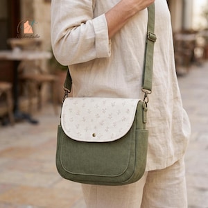 May include: A crossbody bag featuring a solid olive green body and a cream-coloured flap with a delicate, repeating floral pattern. The bag includes an adjustable olive green shoulder strap, side pockets, and a single metallic snap closure.