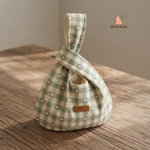 May include: A sage green and white gingham fabric bag with a small heart pattern throughout. The bag features a Japanese-style knot handle design and a rectangular brown leather tag with the word HANDMADE embossed on the front surface.