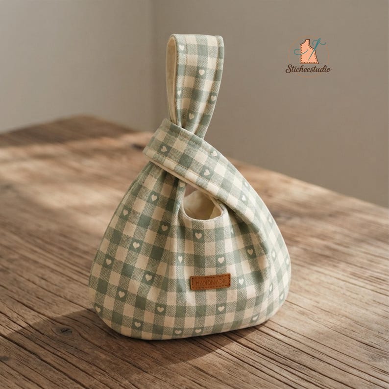 May include: A sage green and white gingham fabric bag with a small heart pattern throughout. The bag features a Japanese-style knot handle design and a rectangular brown leather tag with the word HANDMADE embossed on the front surface.