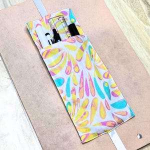 Könnte beinhalten: Ein buntes Federmäppchen aus Stoff mit einem Muster aus weißen, gelben, rosa und blauen Tropfen. Es enthält ein Lineal, Stifte und Marker. Ein weißer Riemen ist befestigt.