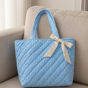 Pode incluir: Uma bolsa tote acolchoada com um padrão de riscas verticais em azul claro e branco. A bolsa tem duas alças de tecido a condizer e está decorada com uma fita de cor creme atada num laço no lado superior direito do painel frontal.