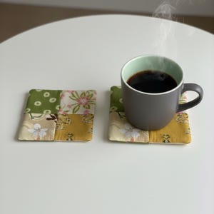 Könnte beinhalten: Eine graue Tasse mit dunklem Kaffee steht auf einem Patchwork-Untersetzer. Der Untersetzer besteht aus Stoffquadraten in Grün-, Gelb- und Cremetönen mit floralen Mustern. Ein zweiter Untersetzer befindet sich neben der Tasse. Dampf steigt vom Kaffee auf.