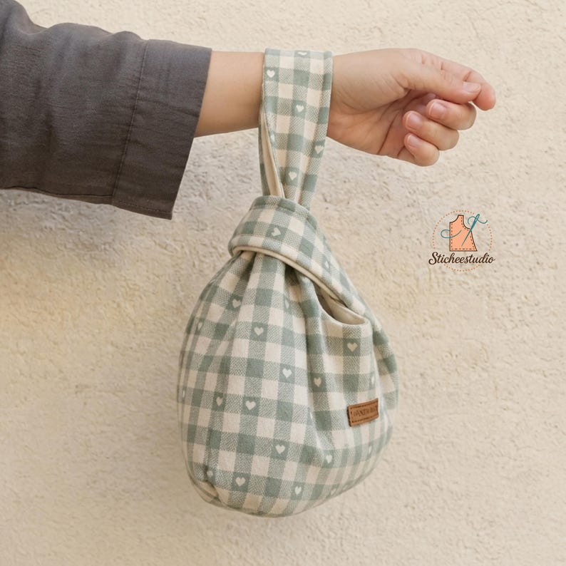 May include: A fabric knot bag featuring a sage green and cream gingham check pattern with small white heart icons throughout. The bag has a single integrated handle loop and a small rectangular brown leather-like tag on the side.