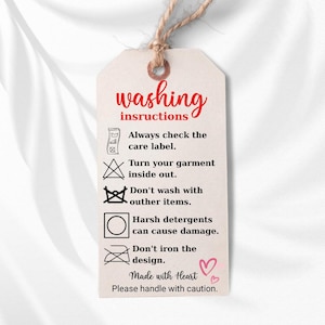 a white tag with instructions for washing clothes, including checking the care label, turning the garment inside out, avoiding harsh detergents, and not ironing the design.