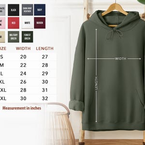 a green hoodie hanging on a hanger, with a size chart and measurements provided. The chart lists the size options and corresponding measurements, such as S, M, L, XL, 2XL, and 3XL, along with the corresponding widths. The image also includes a plant in the background, adding a natural element to the scene.