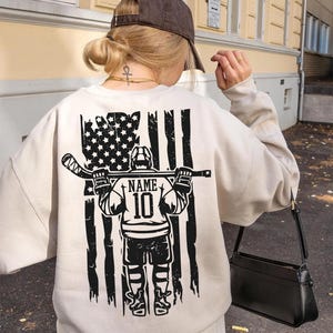 A person wearing a white sweatshirt with a graphic design on the back, including an American flag and a hockey player.