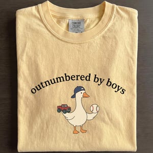 A yellow t-shirt with a cartoon goose holding a baseball and a truck, along with the text "outnumbered by boys" printed on it.