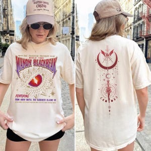 a woman wearing a white t-shirt with a graphic design and text on it. The woman is standing in front of a city street with buildings in the background.