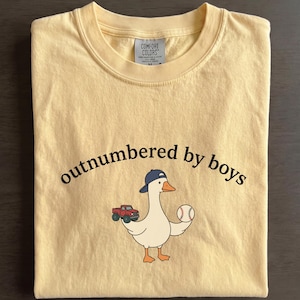 A yellow t-shirt with a cartoon goose holding a baseball and a truck, along with the text "outnumbered by boys" printed on it.