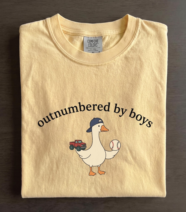 A yellow t-shirt with a cartoon goose holding a baseball and a truck, along with the text "outnumbered by boys" printed on it.