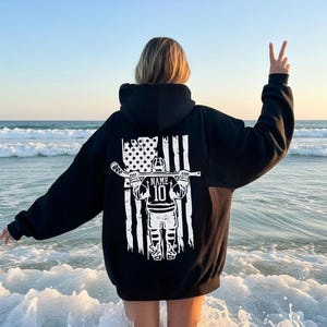 A person standing in the ocean, wearing a black hoodie with a graphic design on the back, making a peace sign with their hand.