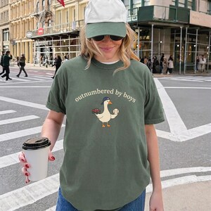 A woman wearing a green t-shirt with a baseball graphic and a cap is holding a coffee cup and standing on a city street.