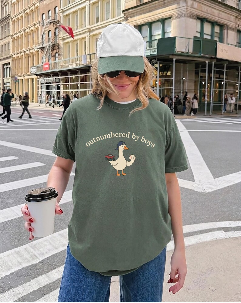 A woman wearing a green t-shirt with a baseball graphic and a cap is holding a coffee cup and standing on a city street.