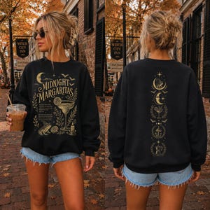 May include: A black long-sleeve sweatshirt featuring a gold graphic print. The front displays the text "MIDNIGHT MARGARITAS" with a margarita glass, a frog, a crescent moon, and stars. The back features a vertical column of celestial and botanical icons.