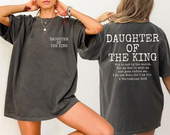 May include: Dark gray t-shirt with the text "DAUGHTER OF THE KING" on the front and back. The back of the shirt also features a Bible verse. The shirt has short sleeves and a relaxed fit.