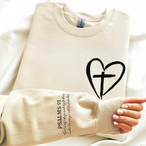 May include: Cream-colored sweatshirt with a black heart and cross design. The sleeve features the text "PSALMS 91" and a Bible verse. The sweatshirt is made of a soft material.