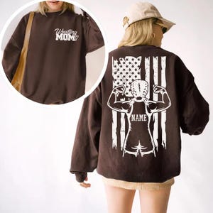 A woman wearing a brown sweatshirt with a graphic design on the back, featuring a muscular figure and an American flag.