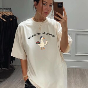 A woman is taking a selfie in a clothing store, wearing a white t-shirt with a cartoon duck and the text "outnumbered by boys".