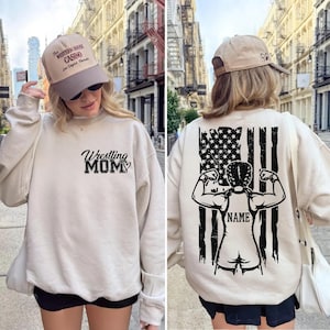 A woman wearing a white sweatshirt with a wrestling theme and a hat, standing in front of a cityscape.