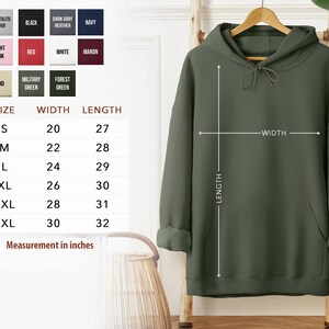 a green hoodie hanging on a hanger, with a size chart and measurements provided. The chart lists the size options and corresponding measurements, such as S, M, L, XL, 2XL, and 3XL, along with the corresponding widths. The image also includes a plant in the background, adding a natural element to the scene.