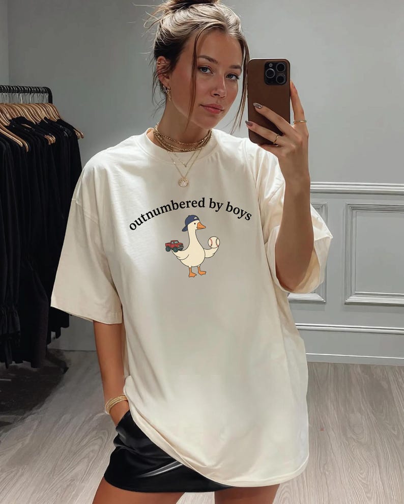 A woman is taking a selfie in a clothing store, wearing a white t-shirt with a cartoon duck and the text "outnumbered by boys".
