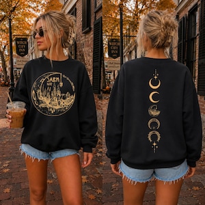 May include: A black long-sleeve sweatshirt featuring a gold graphic on the front of a crescent moon containing the text Salem 1692 with candles and crystals. The back displays a vertical sequence of moon phases and celestial symbols in gold.