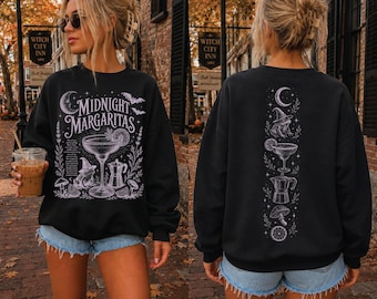 May include: A black crewneck sweatshirt featuring a white graphic print. The front displays the text Midnight Margaritas with a cocktail glass, a frog in a witch hat, and botanical elements. The back shows a vertical design of these same icons.