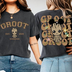 May include: A charcoal gray t-shirt featuring a graphic of Groot on the front with the text GROOT EST. 2014 GUARDIANS. The back displays a larger Groot graphic with mouse ears, surrounded by repeating GROOT text and small icons in tan and green.