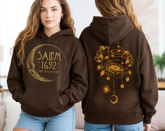 May include: A brown hooded sweatshirt with a front pocket and a gold graphic. The front graphic says "Salem 1692" inside a crescent moon, with the text "They Missed One" below. The back features a gold celestial design.