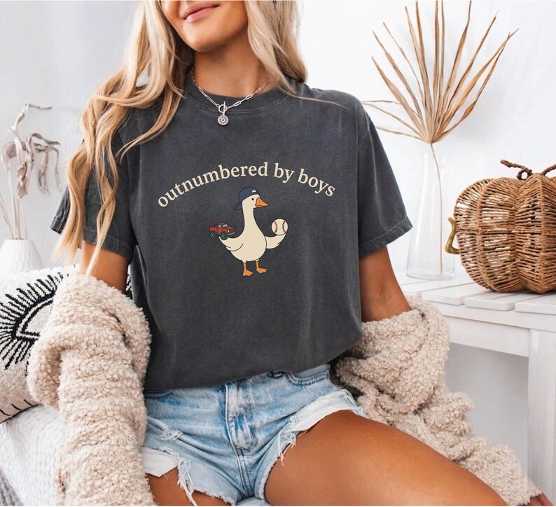 A woman wearing a black t-shirt with a cartoon goose holding a baseball and the text "outnumbered by boys" is sitting in a room with a wicker basket and a plant in the background.