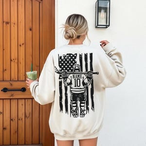 A woman wearing a white sweatshirt with a graphic design of a hockey player holding a hockey stick and an American flag on the back.