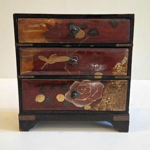 May include: A small, three-drawer wooden chest with a black frame and legs. The drawers are a deep red with gold floral and bird designs. The chest is likely vintage or antique.