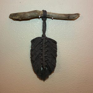 May include: A dark grey macrame feather wall hanging suspended from a natural piece of driftwood. The fibre art piece features intricate knotted cord patterns forming a leaf shape, with a central spine and frayed edges hanging from a wooden branch.