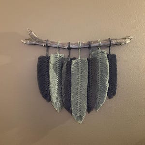 May include: A macrame wall hanging featuring a natural driftwood branch with six gray and blue-gray feather-shaped macrame pieces suspended below. The feathers are arranged in a gradient of colors, creating a textured, decorative wall art piece.