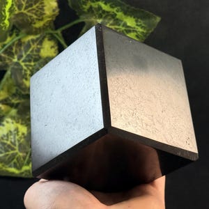 Puede incluir: Un objeto oscuro y cuadrado con una superficie texturizada de color claro y bordes oscuros. El cubo se sostiene en una mano, con hojas verdes y amarillas en el fondo. El objeto parece ser un artículo decorativo.