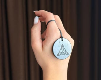Puede incluir: Un colgante redondo gris con un diseño de nudo celta, suspendido de un cordón negro. El colgante se muestra sobre un fondo marrón.