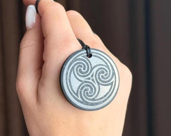 Puede incluir: Un colgante circular gris con un diseño de Triskel celta, borde negro y cordón negro. El símbolo del Triskel presenta tres espirales entrelazadas. El colgante está sostenido por una mano.