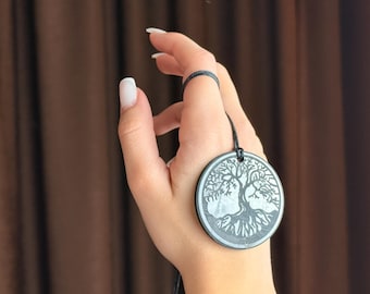 Puede incluir: Colgante redondo plateado con un diseño detallado de árbol de la vida. Cuelga de un cordón negro. El fondo es borroso y marrón. El diseño es intrincado.